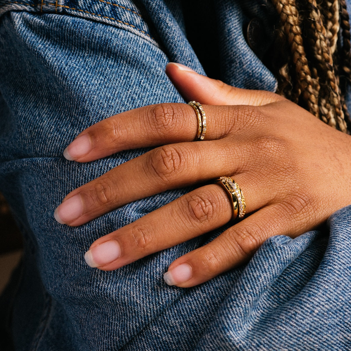 A hand with neatly manicured nails wears the Regency Stacking Ring, a yellow gold plated band, resting on a denim jacket sleeve with braided hair in the background.