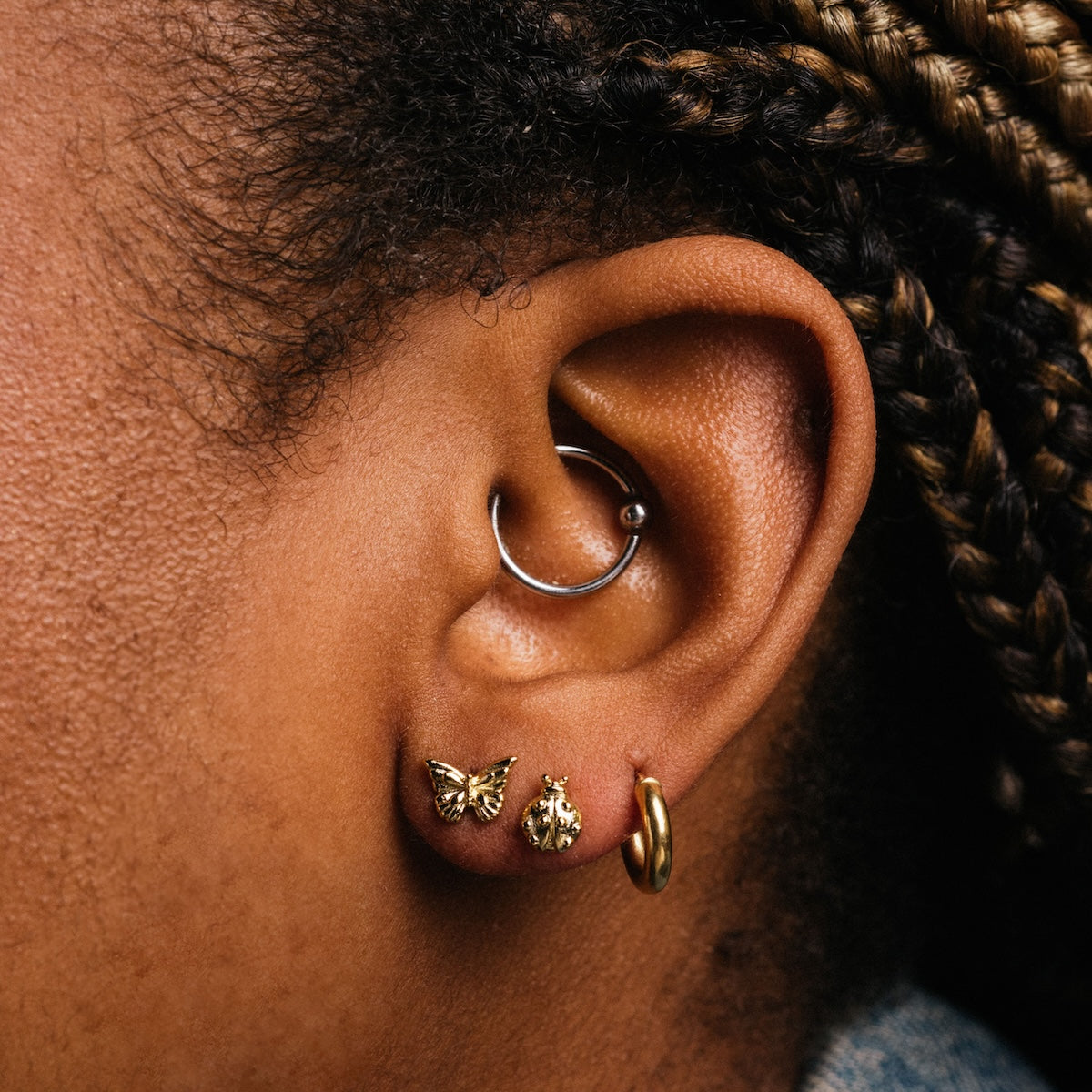 Close-up of a person's ear with four piercings: a minimal 14k gold plated butterfly earring, a round stud, a small hoop in the lobe, and a silver hoop in the cartilage. Braided hair and Lady Bug Studs add stylish detail.