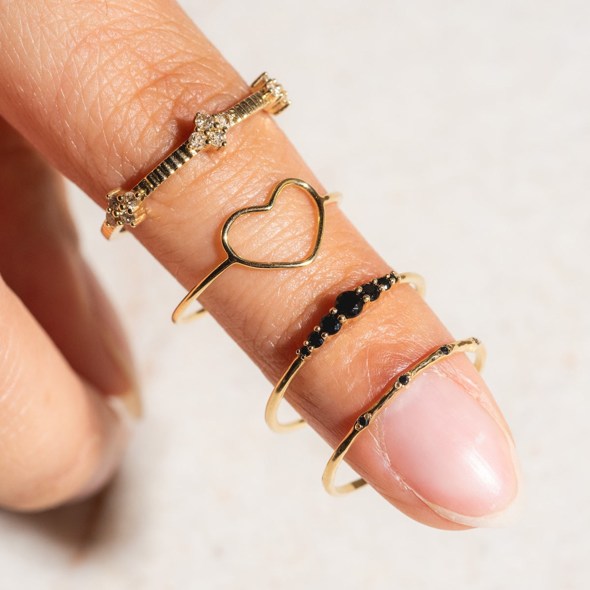 A hand wears four gold rings on the index finger, including the Solid Gold Delicate Heart Ring. The light, neutral background showcases the beauty of Family Gold pieces.