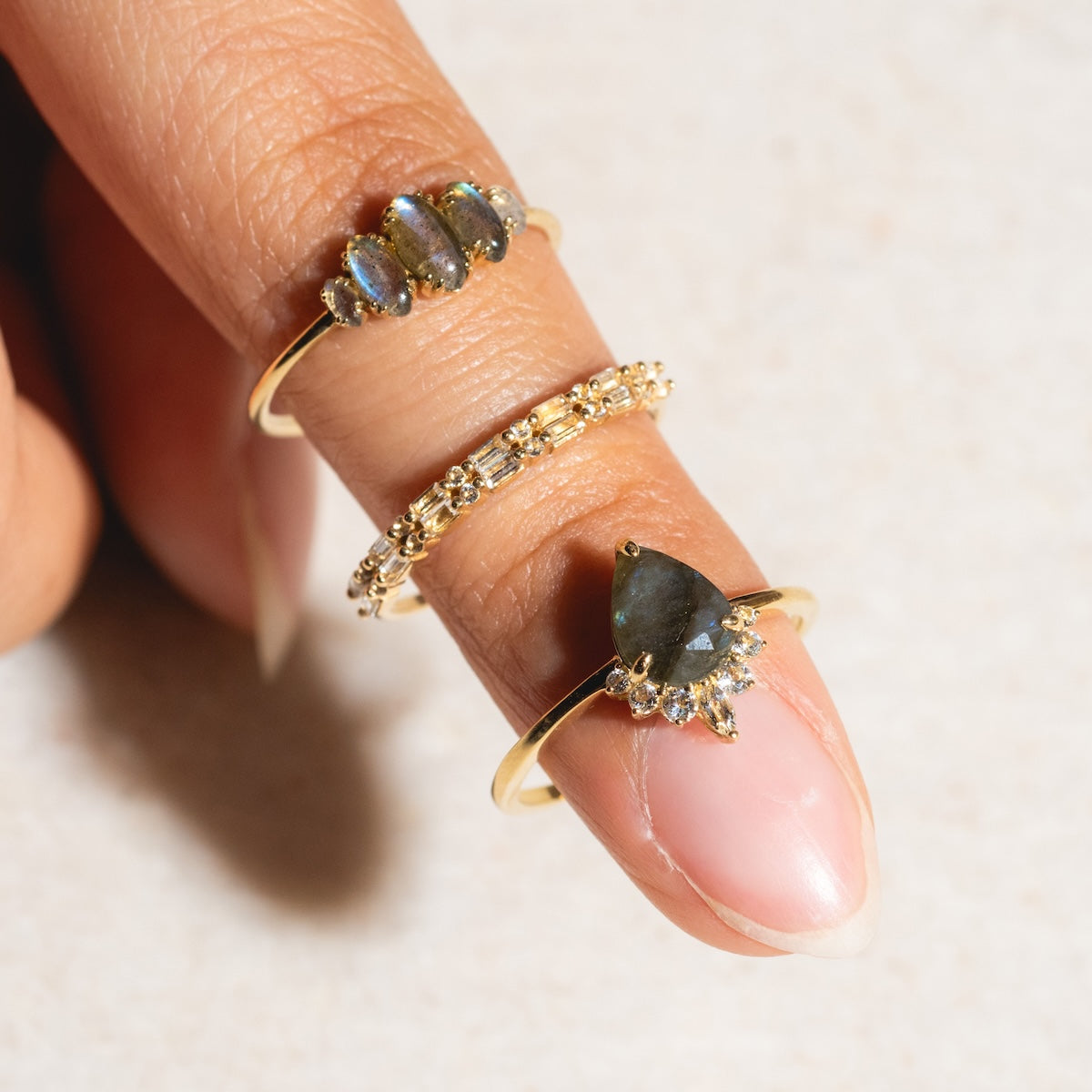 A close-up of a hand with three gold rings: the Solid Gold White Topaz Deco Band, a delicate floral ring, and a pear-shaped dark gemstone ring surrounded by clear stones.