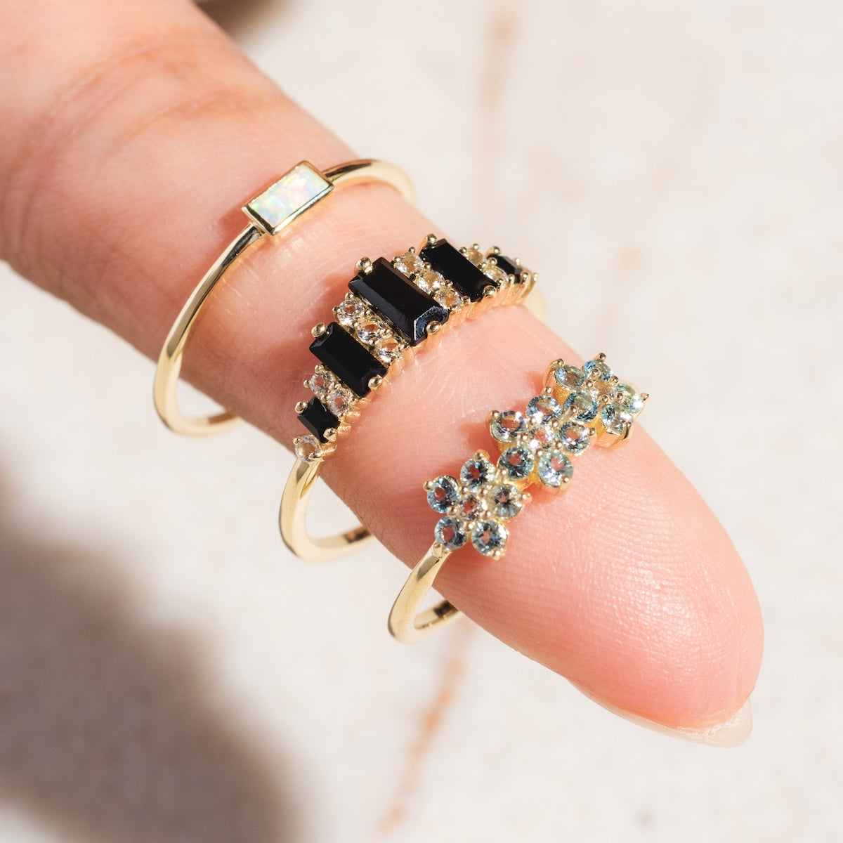 A close-up of three gold rings on a finger: the Solid Gold Delicate Opal Ring with a rectangular stone, one with vertical black stones and clear gems, and a Family Gold ring with sparkling round gemstones.