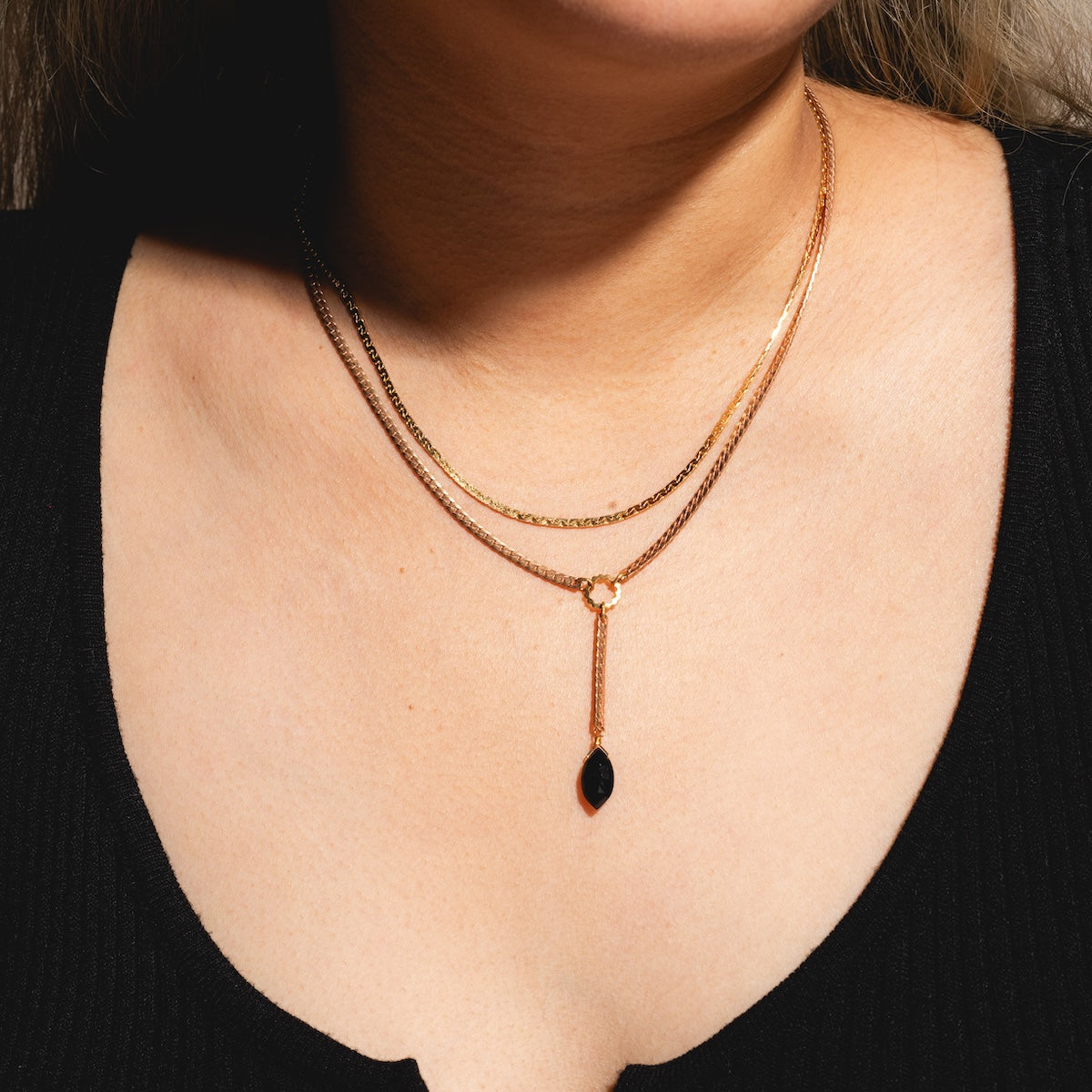 A person is shown from shoulders to collarbone wearing a black ribbed top and layered gold necklaces, including the Palmer Gemstone Necklace with a small black gemstone pendant.
