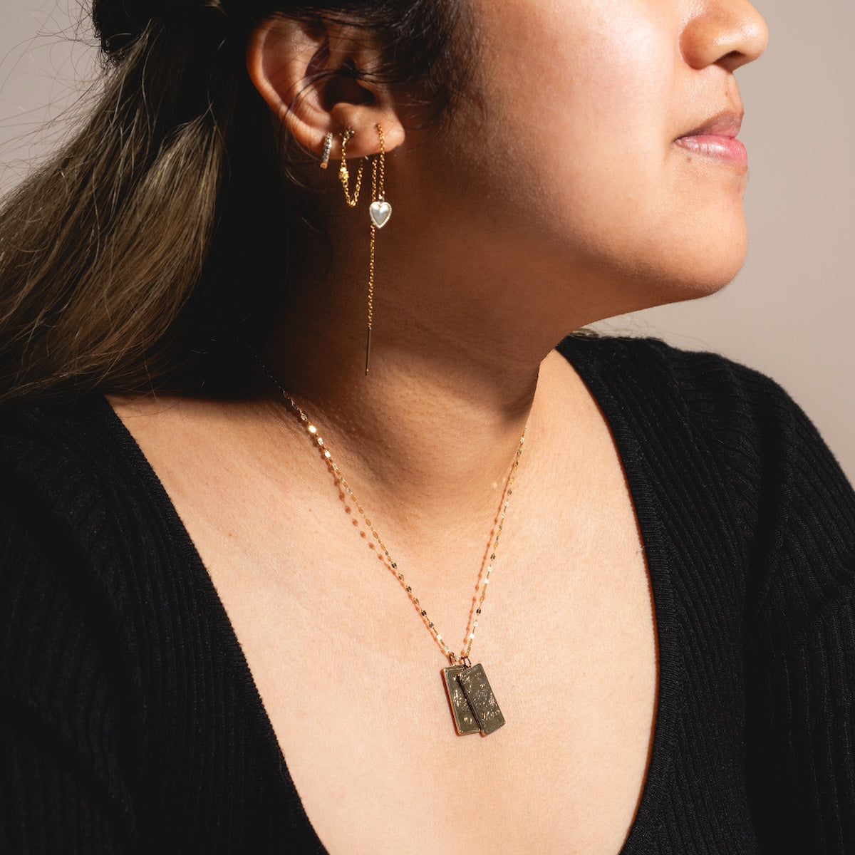 A woman in a black top is shown from the shoulders up. She wears gold hoop earrings with hanging chains and a 14k solid gold Tarot Card Charm, along with a gold necklace featuring two rectangular pendants. Her hair is pulled back.