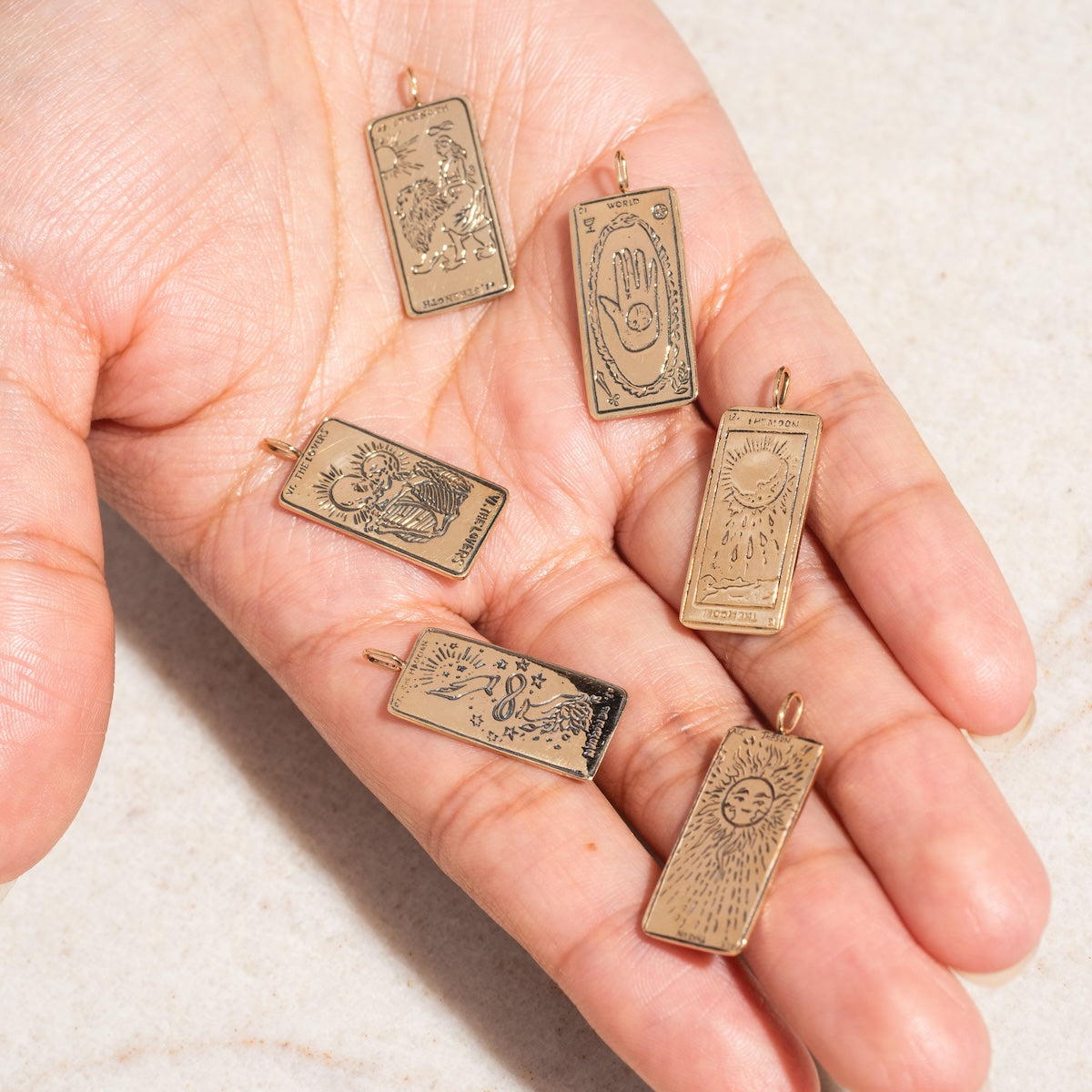 A hand holds six rectangular Tarot Card Charms in 14k solid gold, each engraved with mystical designs—sun, moon, stars, a hand, an eye, and animals—displayed on a light surface; ideal spiritual jewelry or charms.