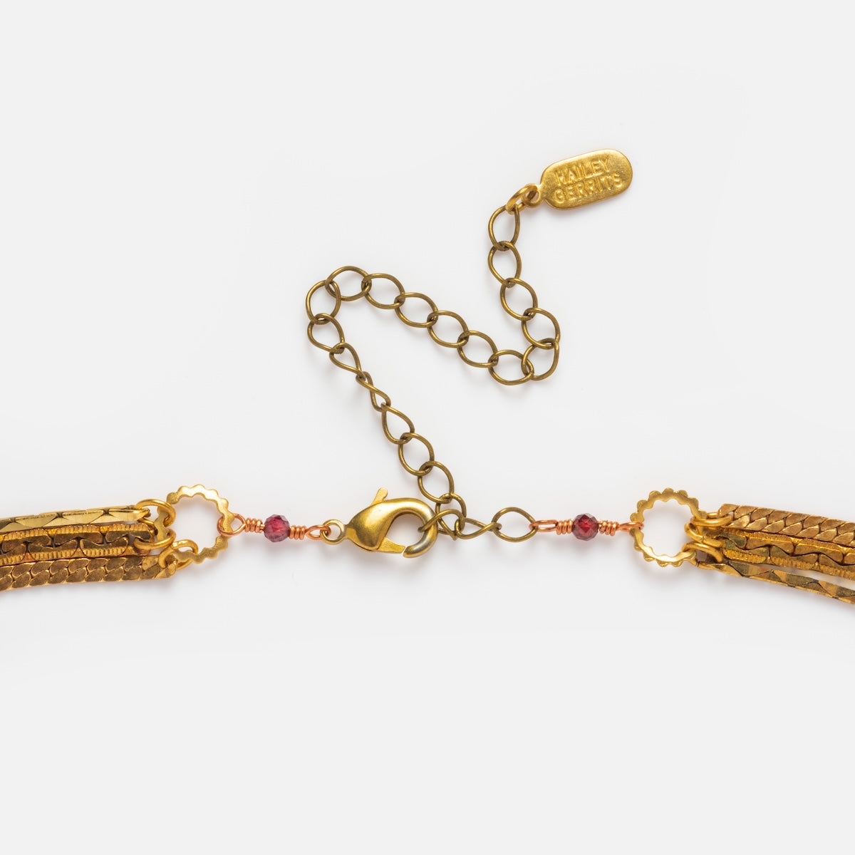 A close-up of the Palmer Gemstone Necklace shows gold layered recycled brass chains with small red garnet beads, a lobster clasp, and an adjustable extender chain featuring a gold tag engraved with text, displayed on a white background.