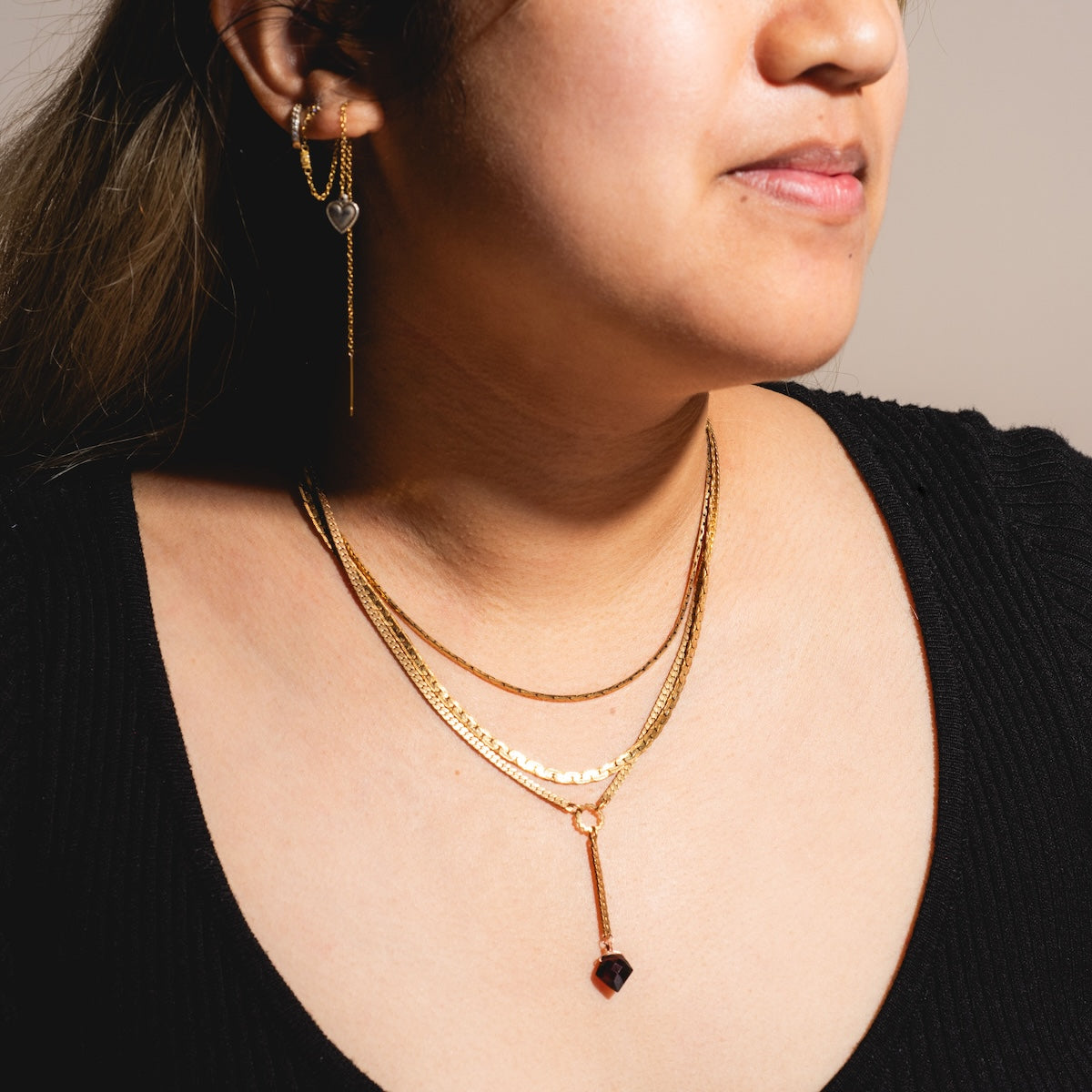 A woman in a black top is shown from the shoulders up, wearing the Palmer Gemstone Necklace with recycled brass chains, garnet accents, and a gold heart-shaped earring.