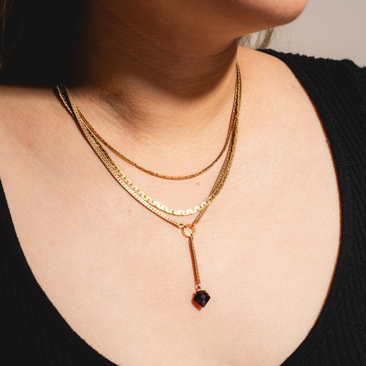 A close-up of a woman in a black top wearing the Palmer Gemstone Necklace—a gold layered chain with one recycled brass chain holding a dark red garnet pendant.