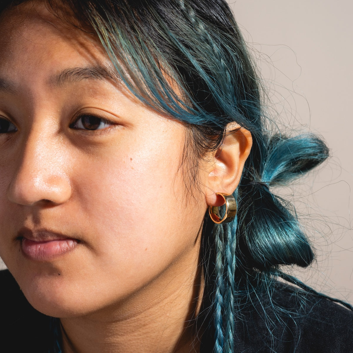 A young person with teal braids and a bun models Solid Gold Flat Chunky Hollow Hoops. The close-up captures their face in soft natural light with a neutral background.