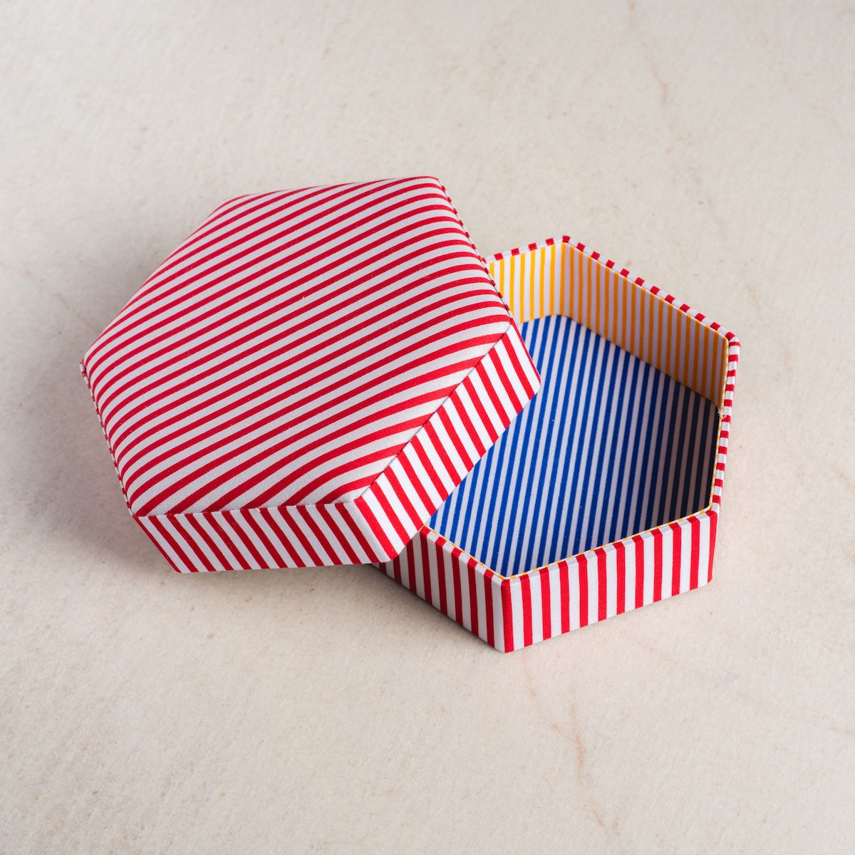 The Pinstriped Jewelry Box features a red and white striped lid, a blue and white striped interior, and yellow-orange striped sides—an ideal, stylish storage solution for jewelry on any light beige surface.