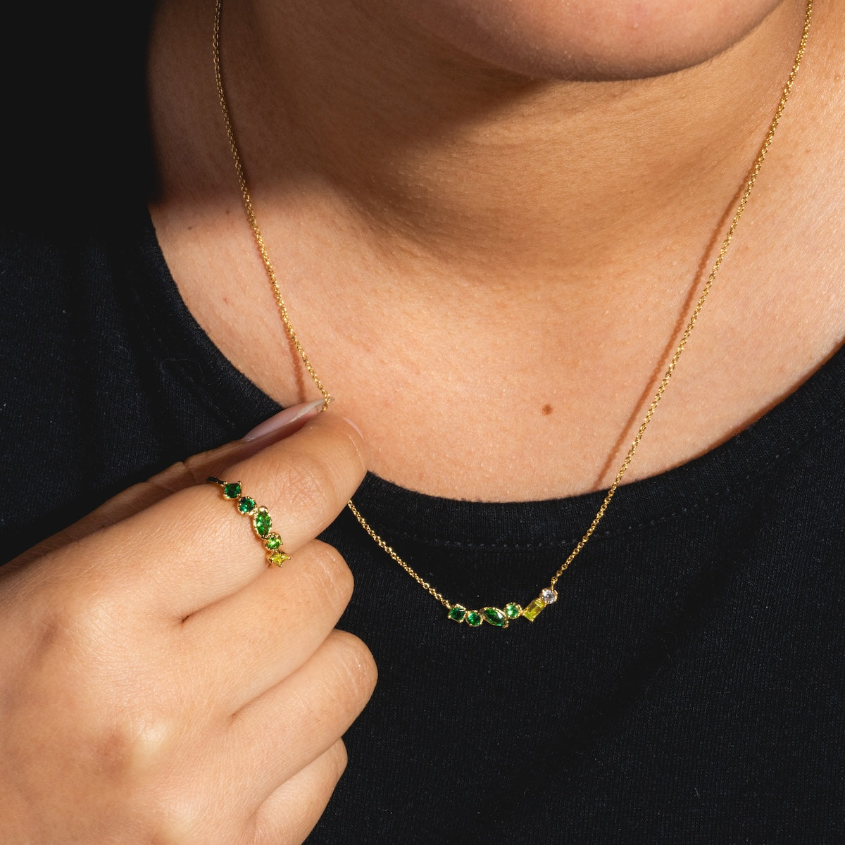 Wearing the Shades of You Ombre Birthstone Ring and Necklace Gift Set, featuring a gold necklace with green and clear gemstones and a matching ring, against a black shirt. The necklace chain is held near the collarbone.