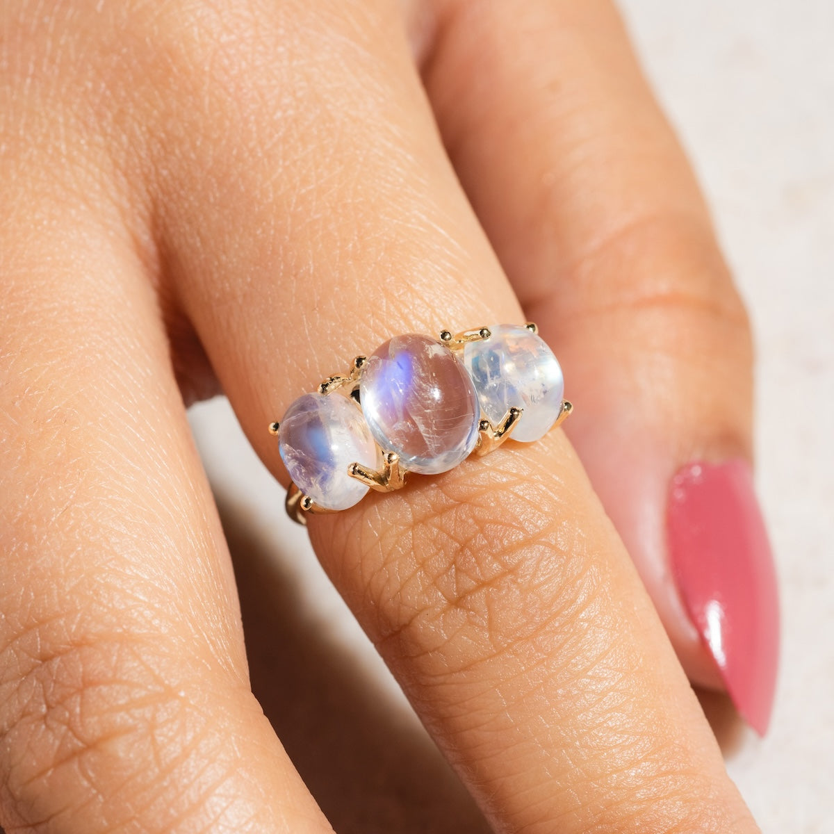 A hand with pink nail polish wears the Sweetest Gumdrop Ring in yellow gold, which features three oval translucent moonstone gems that reflect a subtle blue sheen.