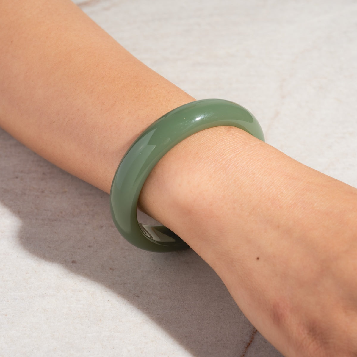 A person’s wrist wears the Smoke Green Jade Quartzite Stone Bangle, its smooth, glossy green stone shown against a light-textured background—a striking jade jewelry piece.
