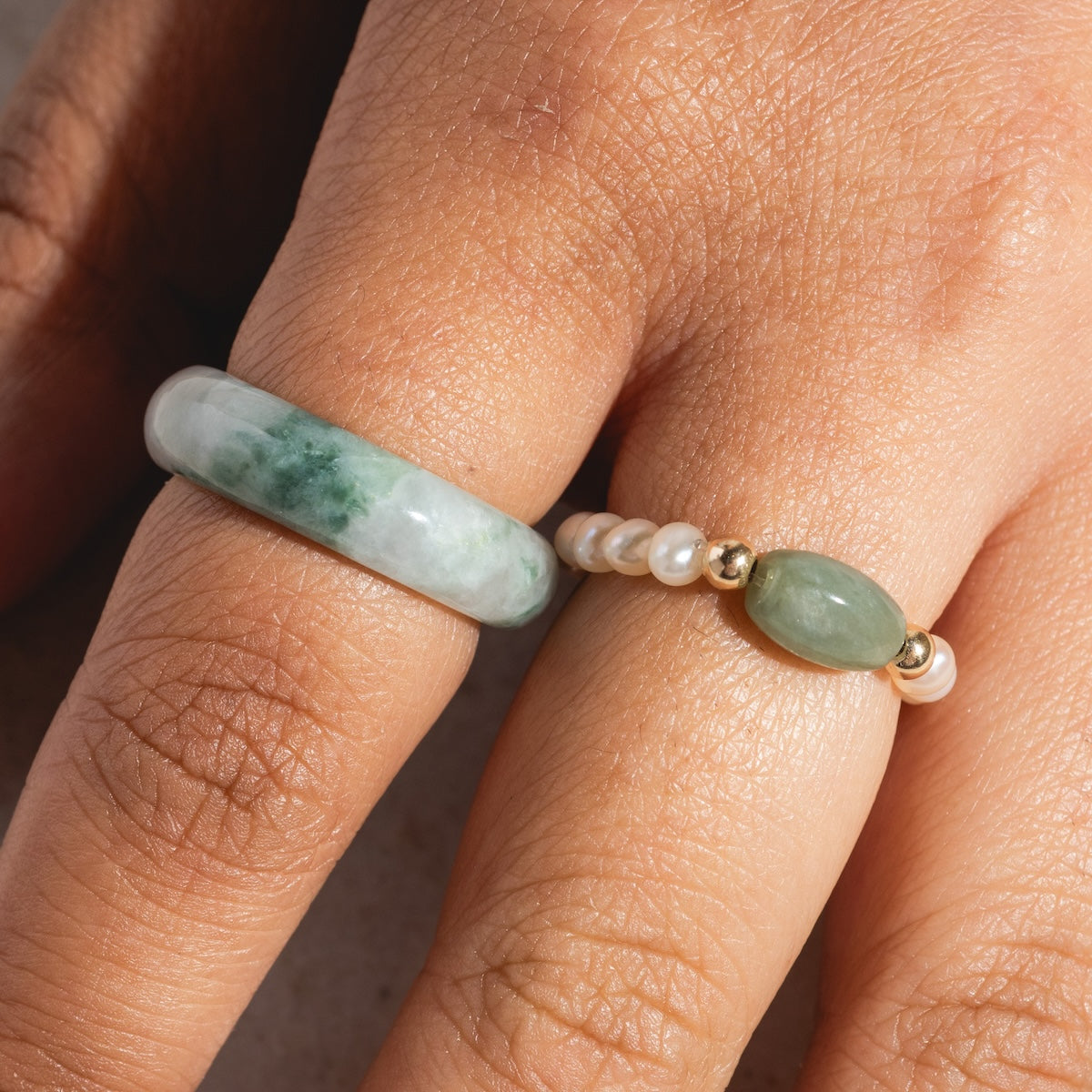 Close-up of two fingers wearing rings; one features the Koi Mottled Green Jade Ring, a thick hand-carved jadeite piece, while the other is a delicate band with small white pearls and a green oval stone.