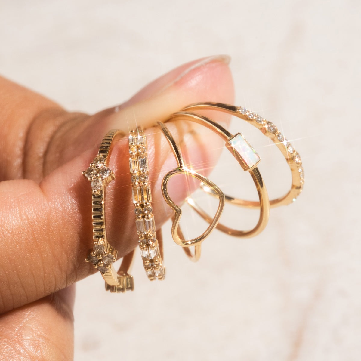A hand displays five delicate gold rings, including the Solid Gold White Topaz Deco Band and vintage-inspired designs with diamonds, a heart shape, and opal—set against a softly blurred background.