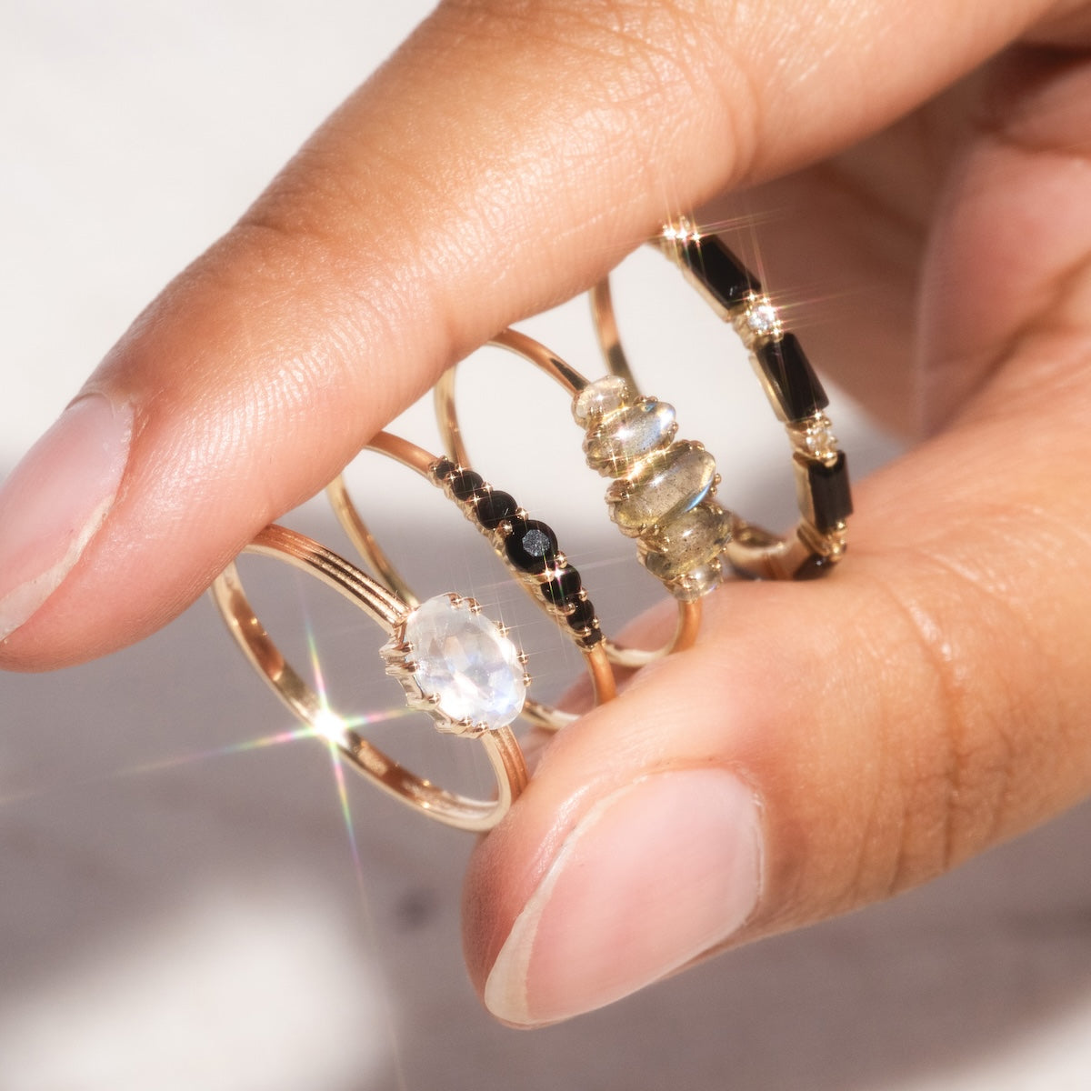 A hand displays four gold rings, including a sparkling Solid Gold Labradorite Marquise Cluster Ring with gemstone accents, stacked white stones, and alternating black and gold bead designs.