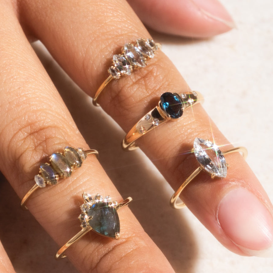 A close-up of a hand with four solid gold rings, including the Solid Gold Labradorite Dew Drop Ring, each featuring a unique gemstone—white topaz, blue stones, and iridescent labradorite—sparkling against a neutral background.