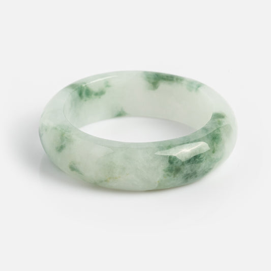 The Koi Mottled Green Jade Ring features a polished, hand-carved jade band with smooth, rounded edges and a blend of light green and white translucent hues, displayed on a plain white background.