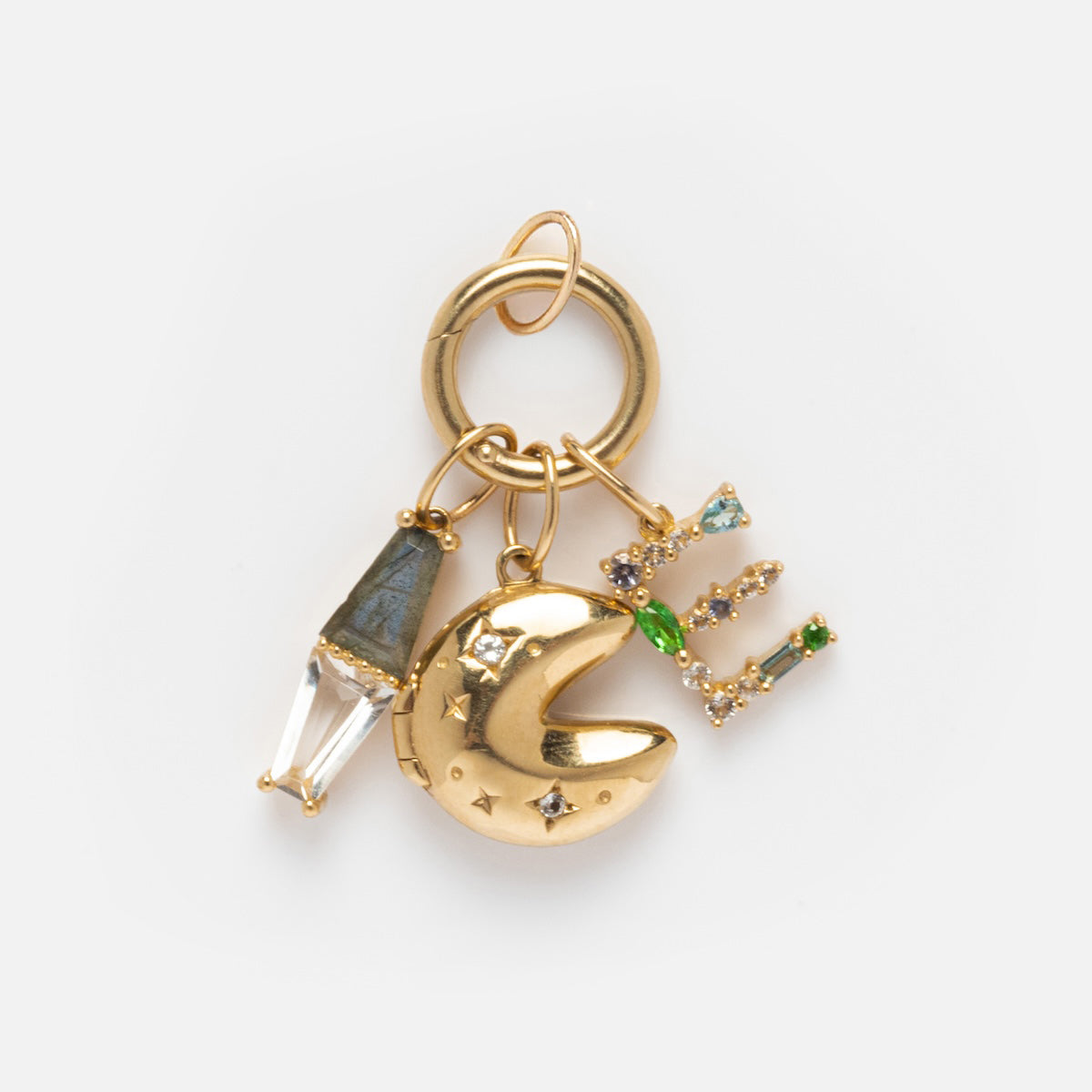 The Solid Gold Charm Clip keyring features three charms: a clear crystal with a gray cap, a gold crescent moon set with gemstones, and a gold letter E embellished with colorful gems. It’s shown on a white background.