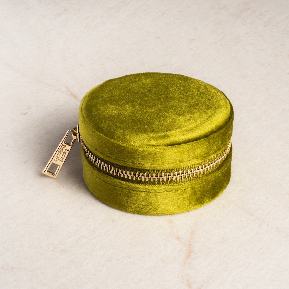 The Weekender Travel Jewelry Case is a small, round, olive-green velvet organizer with a gold zipper and pull, shown on a light beige surface.