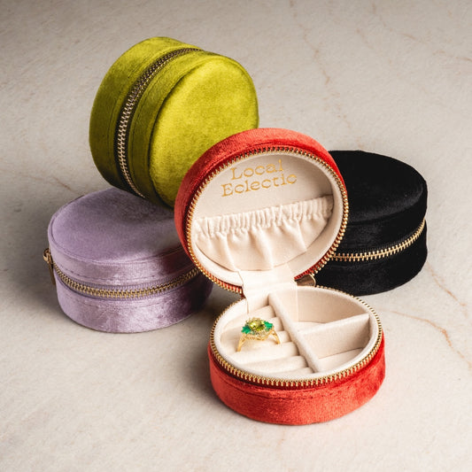 Four round The Weekender Travel Jewelry Cases in green, black, lilac, and red are shown. The open red case displays a gold ring with a green gemstone inside and Local Eclectic stamped in gold on the lids interior.