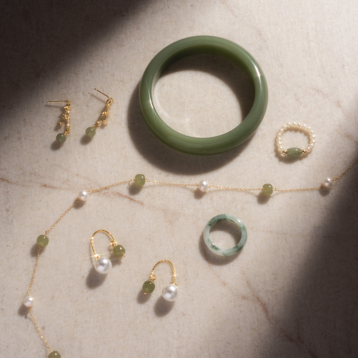 The Koi Mottled Green Jade Ring, two pearl rings, pairs of earrings, and a gold necklace with jade and pearl accents are displayed on a light stone surface with gentle shadows.