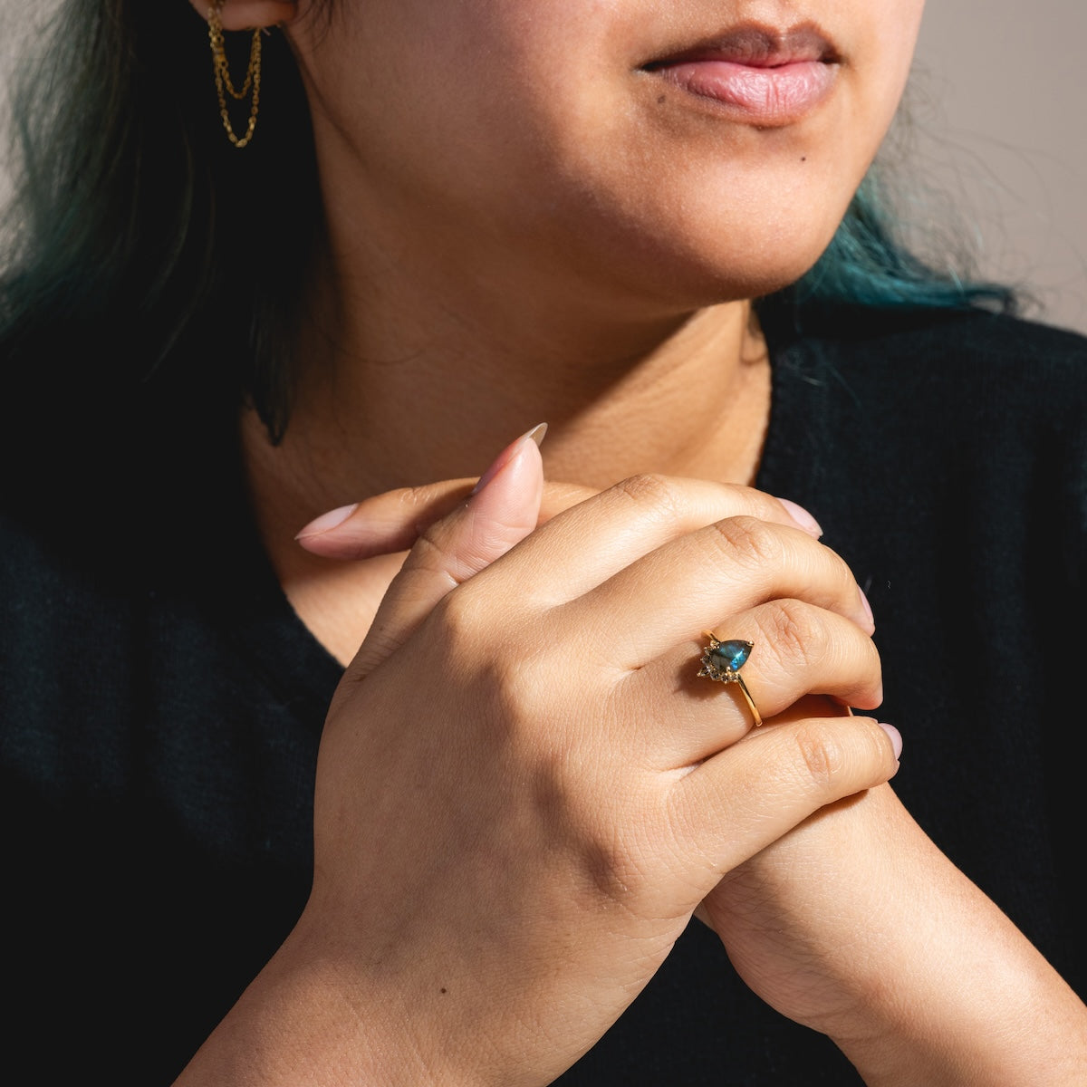 A person with long nails and teal hair clasps their hands, displaying the Solid Gold Labradorite Dew Drop Ring—its blue gemstone catching the light. They wear a black top, gold chain earring, and only part of their face is visible.