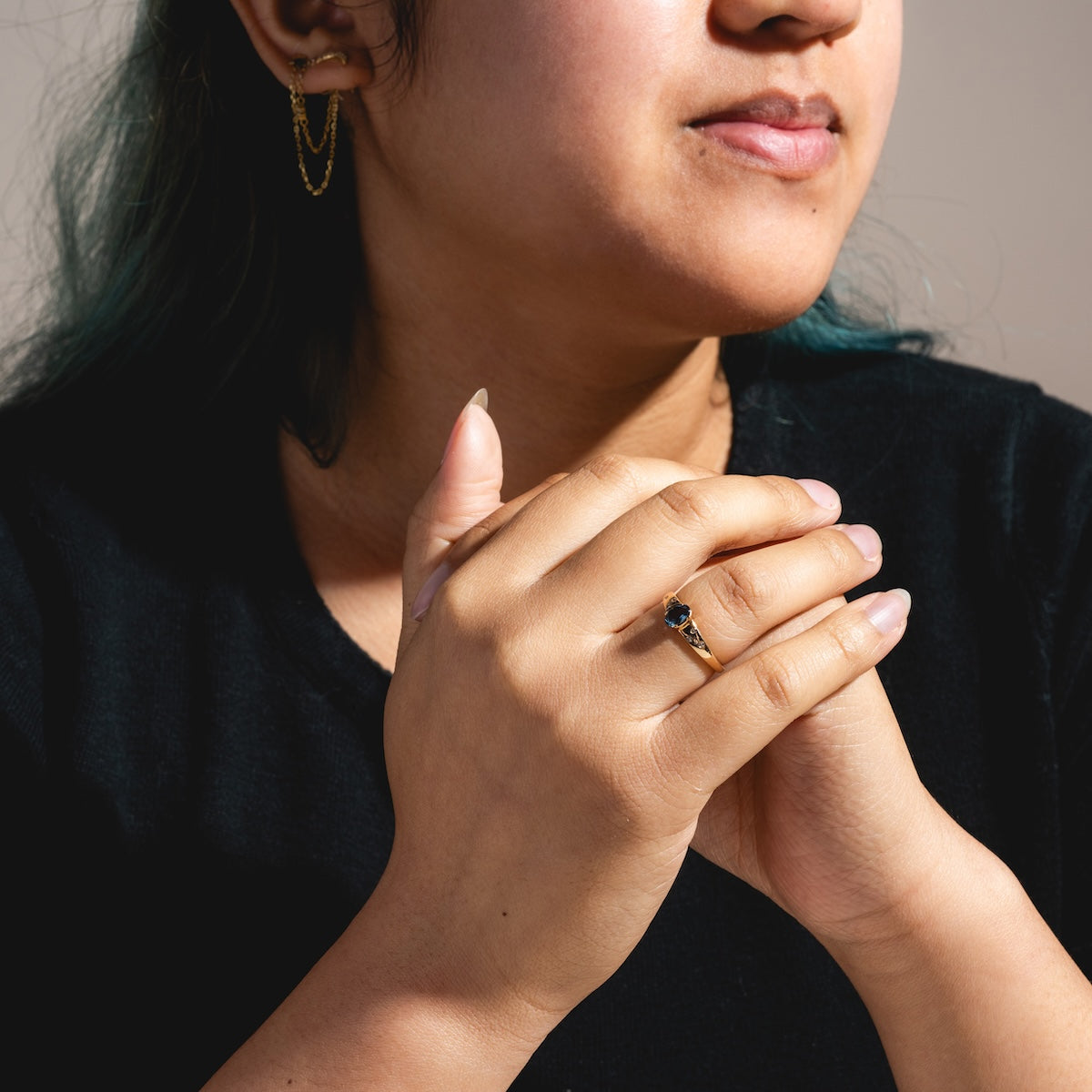 A person in a black shirt clasps their hands, showing off a Solid Gold London Blue Topaz Modern Signet Ring on one finger and gold hoop earrings with chains. Soft lighting creates gentle shadows on their face and hands.