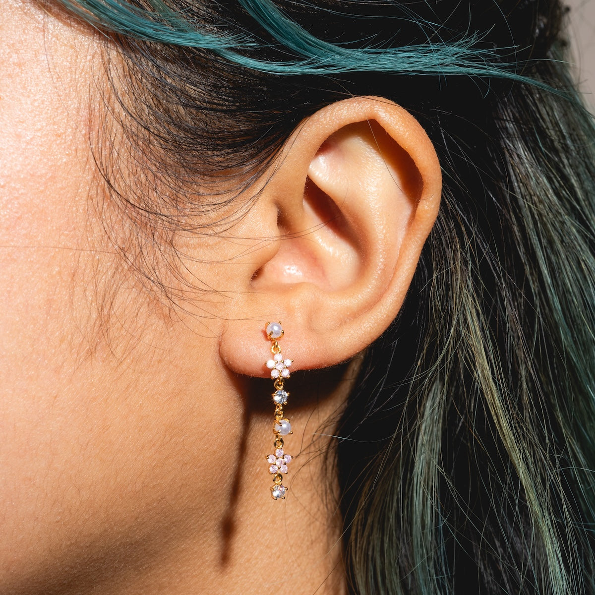 Close-up of a person’s ear wearing the Pearl Petals Dangle Earrings Sample, featuring small flowers and CZ gemstones on 18k yellow gold plated sterling silver posts. The person’s dark hair has teal highlights against a blurred background.