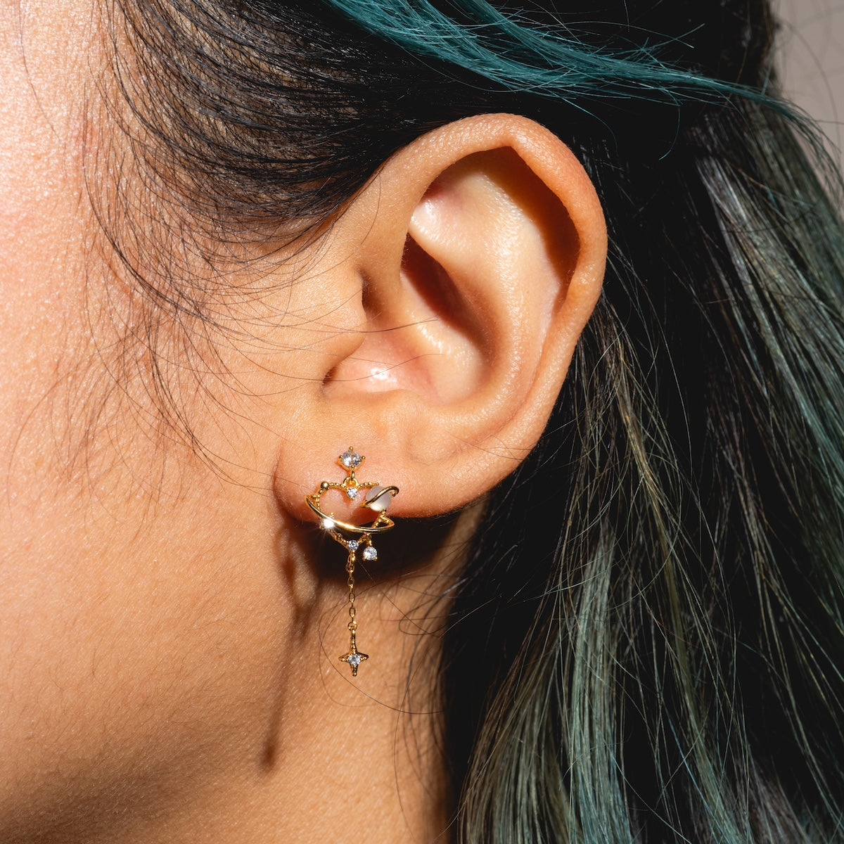 A person with dark hair and green highlights wears the Dream Girl Dangle Hoops Sample—18k yellow gold plated earrings with a planet and star design, dangling chains, and sparkling CZ gemstones.