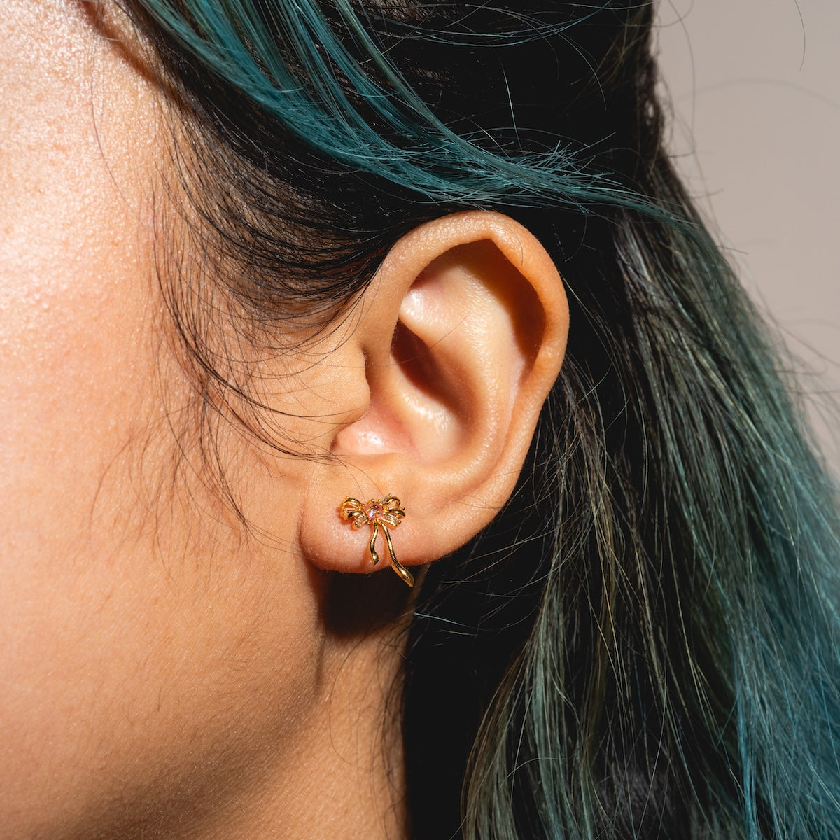 Close-up of a persons ear wearing Coquette Bow Earrings Samples in 18k gold plating with CZ accents. The person’s dark hair has green streaks and is pulled back, set against a soft, neutral background.
