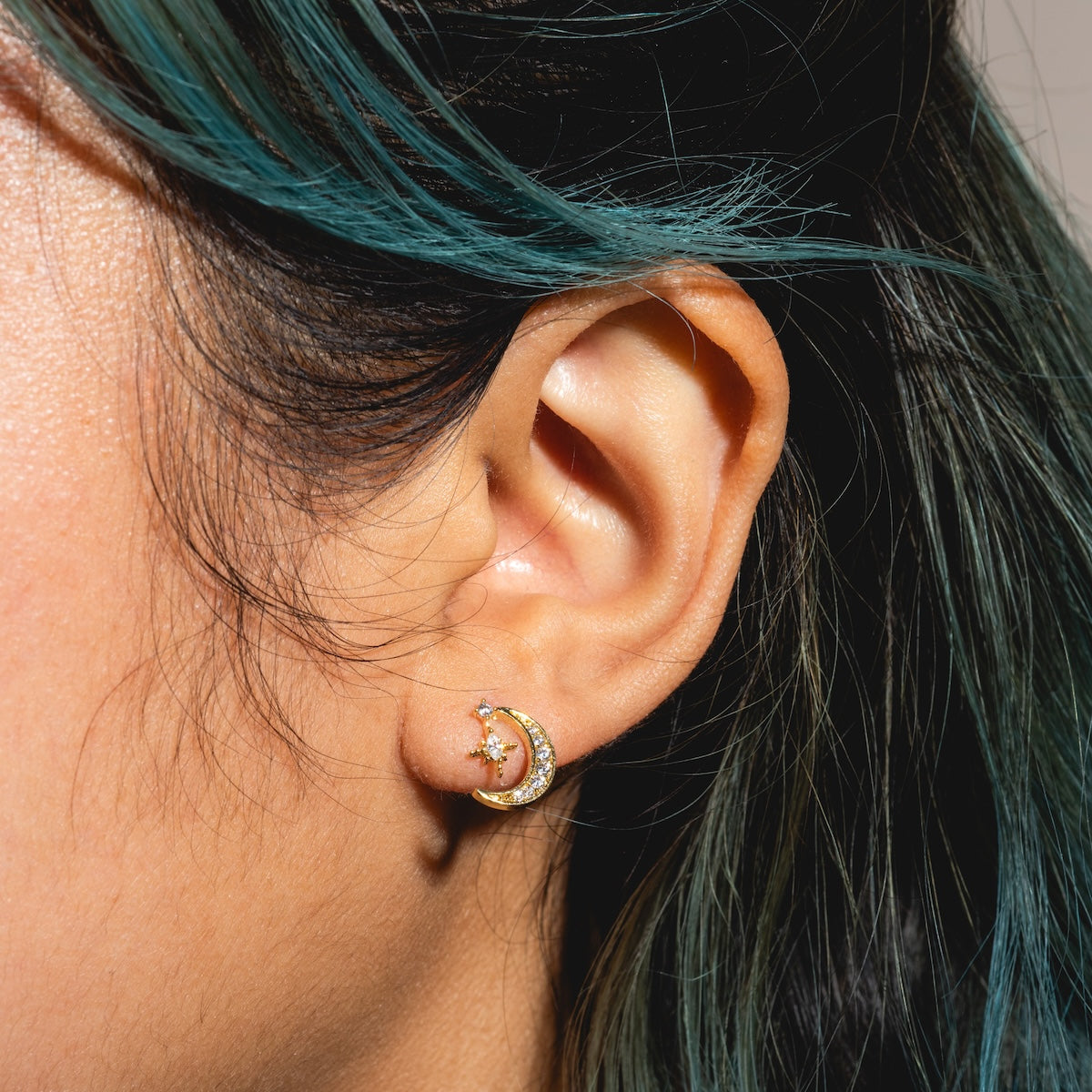 Close-up of a person’s turquoise-tinted hair and ear wearing the Stargazer Moon Earrings Sample: two 18k yellow gold plated earrings—a CZ-set crescent moon and a small stud above, both with sterling silver posts.