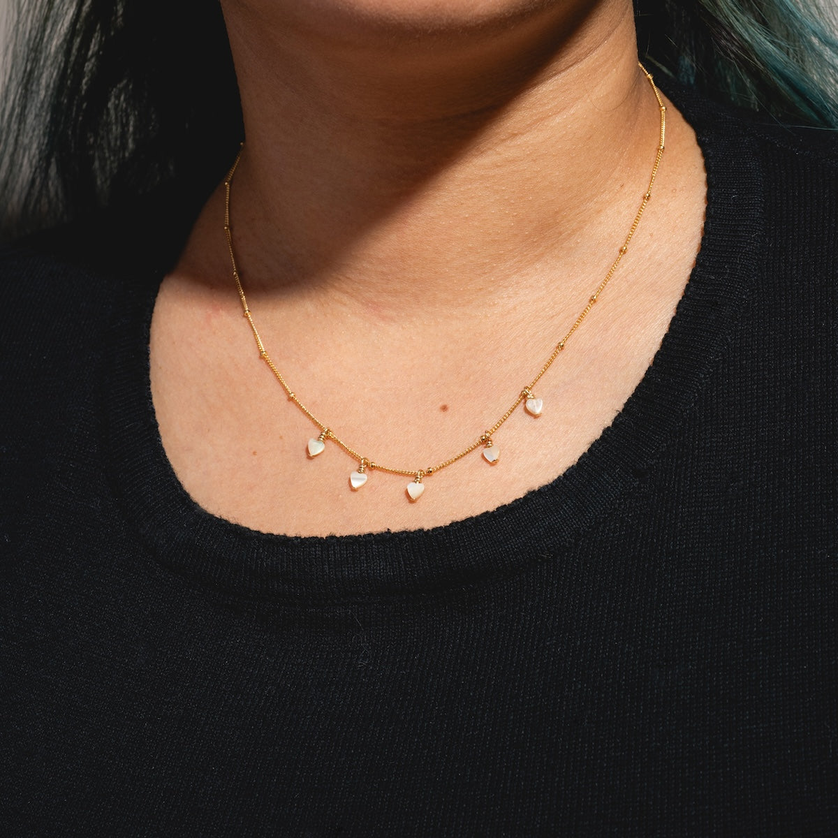 A person wears the Heart Bead Choker Sample—a delicate handmade gold filled chain necklace with small, heart-shaped mother of pearl beads evenly spaced—paired with a black top.
