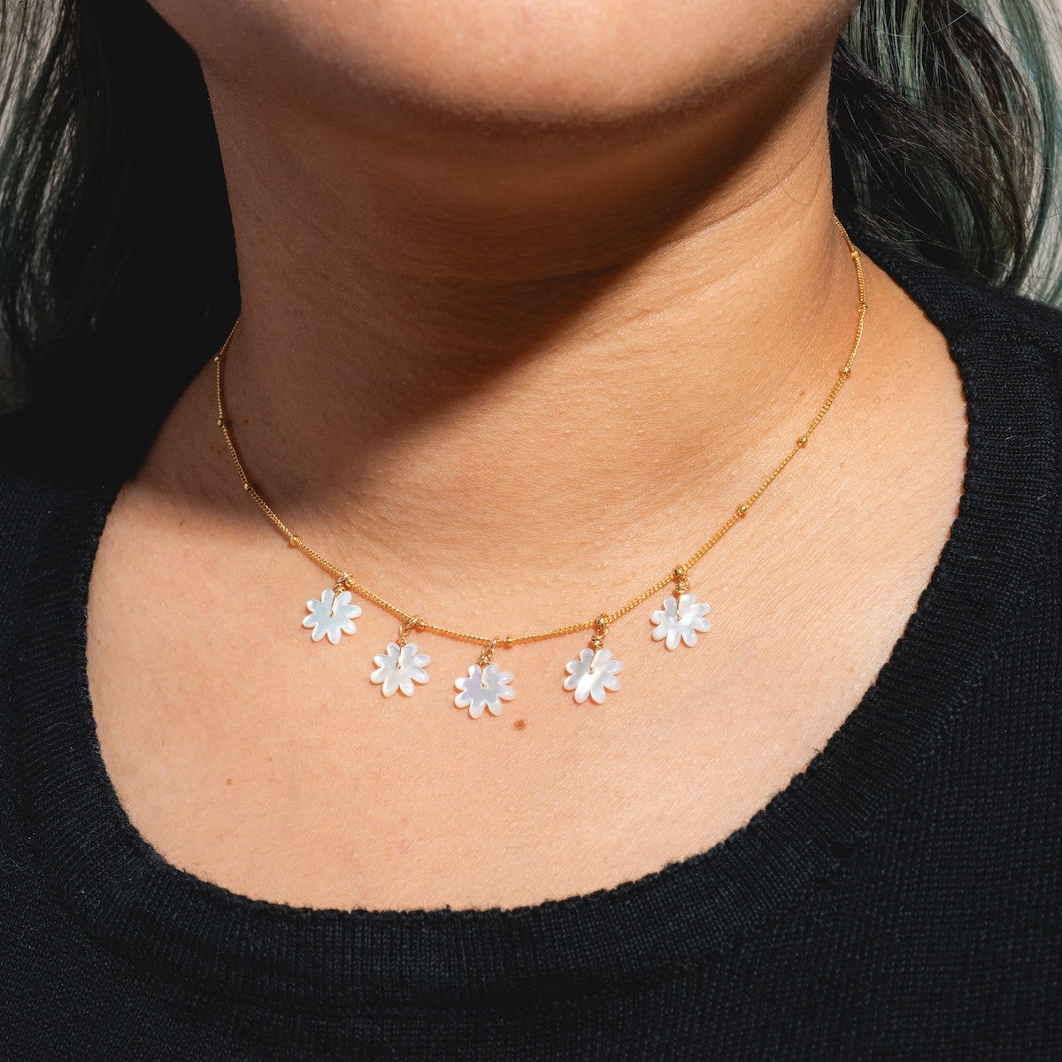 A person wearing the Mother of Pearl Flower Shaker Necklace Sample, featuring five small mother of pearl daisy charms. The individual is in a black top with dark hair partially visible.