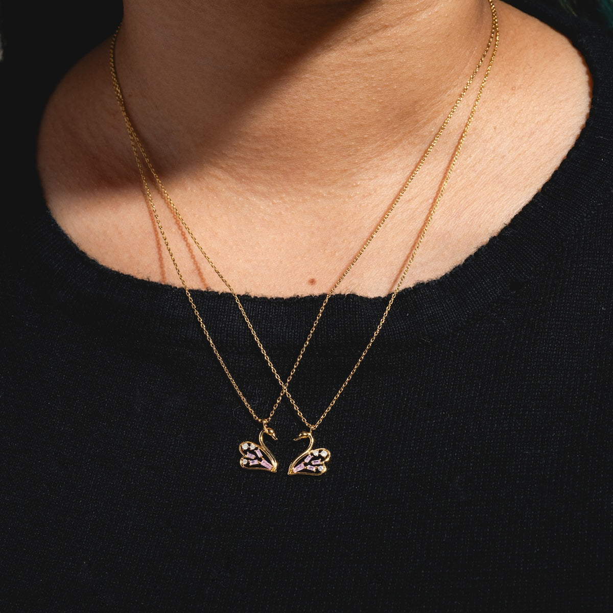 A close-up of a person wearing the Swan Lake Necklace Set Sample—two layered 18k yellow gold plated brass necklaces, each with a swan pendant facing the other over a black sweater for a symmetrical chest design.