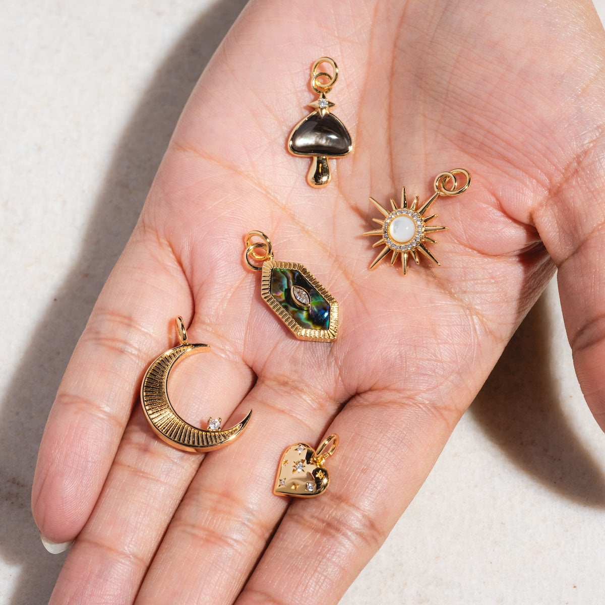 A hand displays five Charms by Elizabeth Stone—14k gold plated celestial pieces: a crescent moon, mushroom, sun with mother of pearl center, faceted gem in gold, and a heart with star—against a light neutral background.