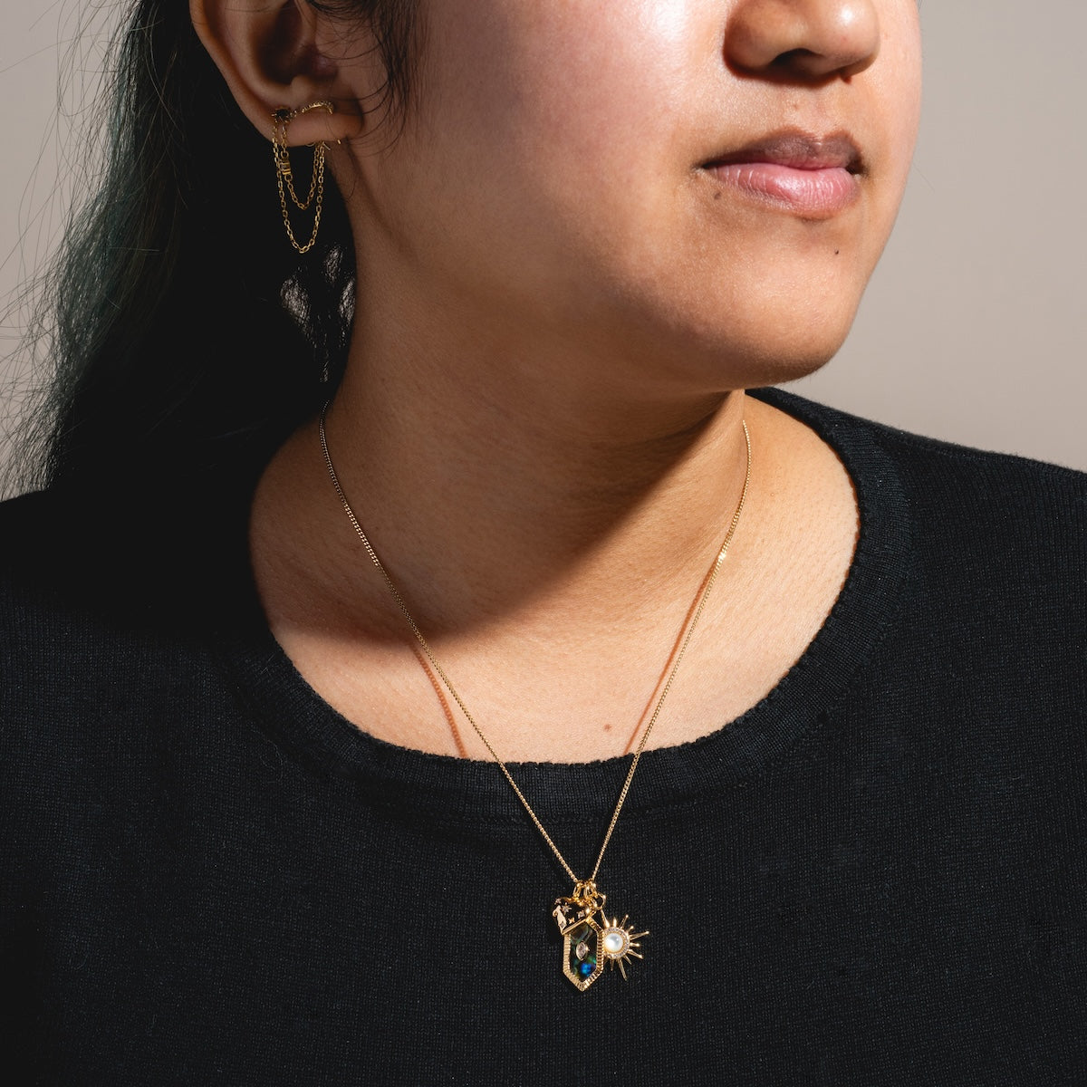 A person wears a black top and Charms by Elizabeth Stone: celestial gold chain earrings with double chains, plus two 14k gold plated necklaces—one with a blue gem pendant, one with a sunburst charm—against a neutral background.