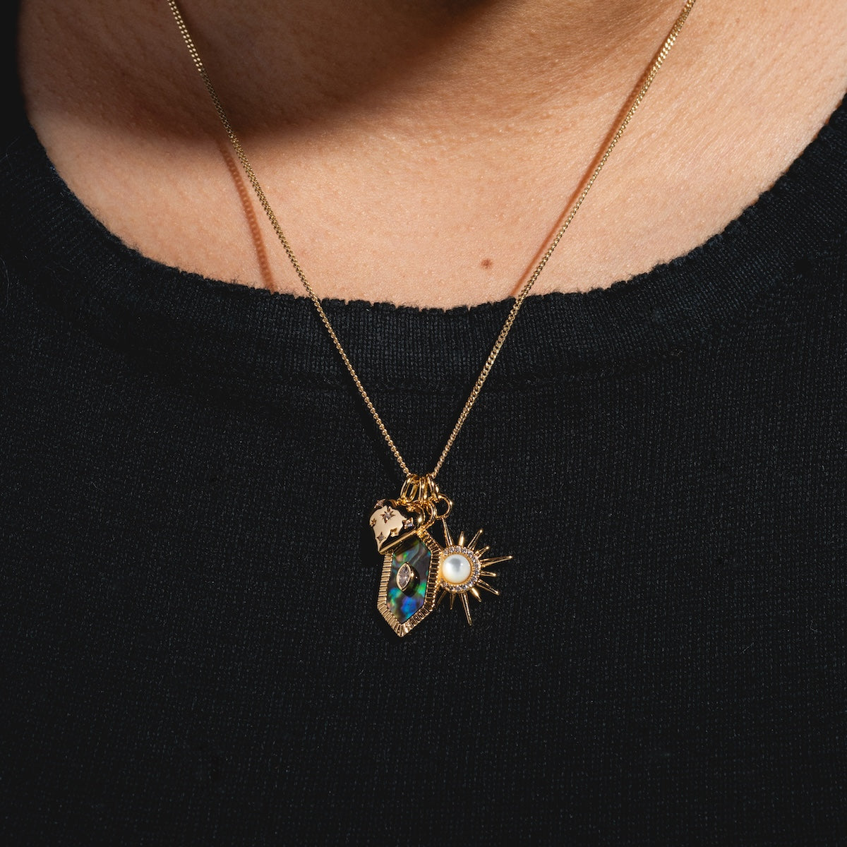 A close-up of the Charms by Elizabeth Stone 14k gold plated necklace, featuring celestial charms—a gem-studded hexagon, a sunburst with mother of pearl center, and a gold charm—worn over a black crew-neck shirt.