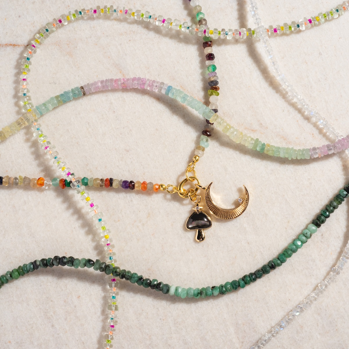 Charms by Elizabeth Stone: Colorful beaded necklaces with green, pink, and clear beads are displayed on a light surface. One necklace features two 14k gold plated celestial charms—a crescent moon and a small mushroom.