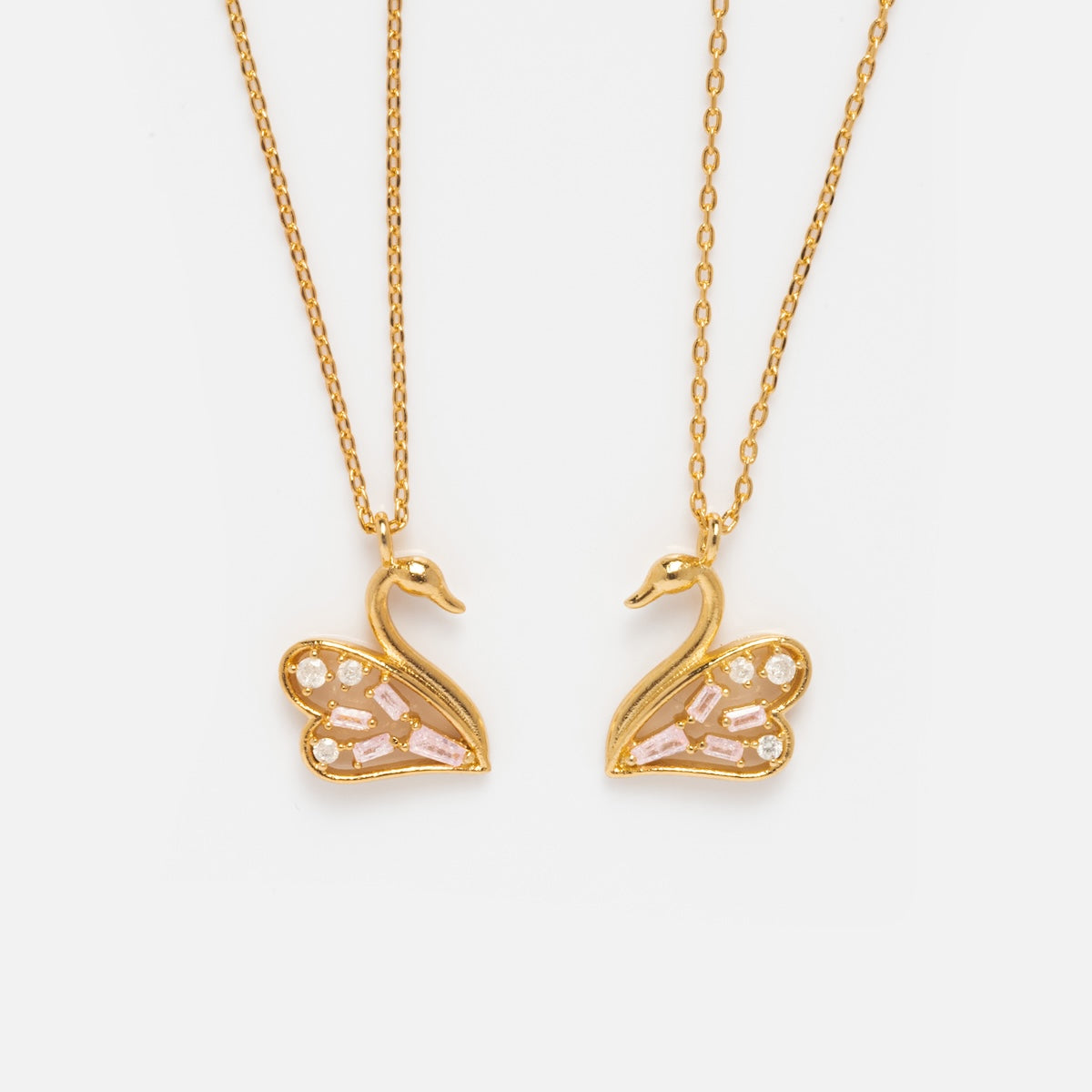 The Swan Lake Necklace Set Sample features two layered necklaces with swan-shaped pendants, crafted in brass and finished with 18k yellow gold plating, showcasing pink and clear gemstone wings against a white background.