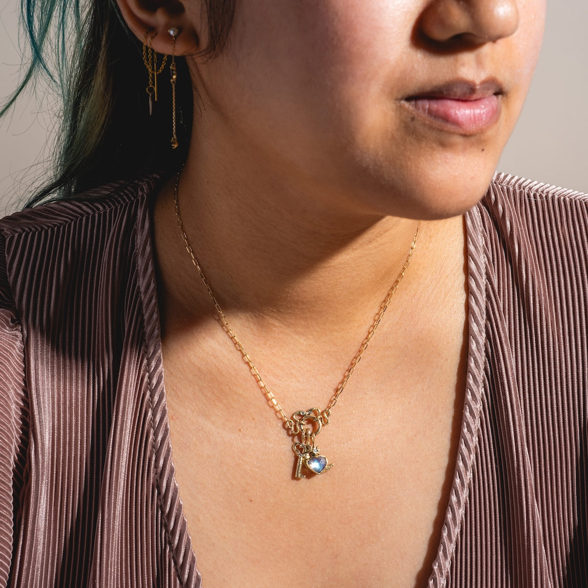 A woman wears the Guided Heart Pendant Charm—14k solid yellow gold heart and key—paired with gold earrings and a pleated mauve V-neck top. The image highlights her lower face, neck, and chest.