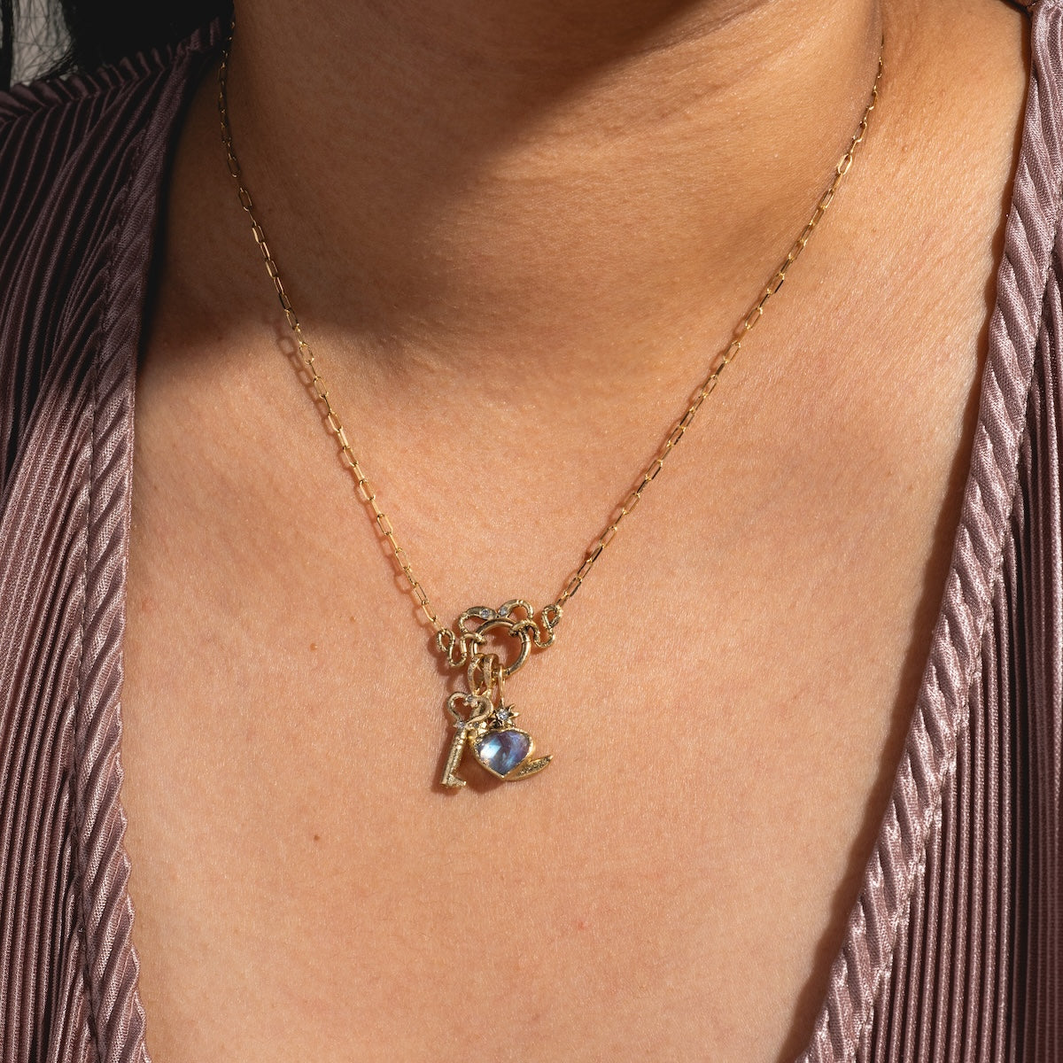 A 14k solid yellow gold chain featuring the Guided Heart Pendant Charm, a key, and a small bird, worn around the neck and paired with a pleated purple V-neck top.