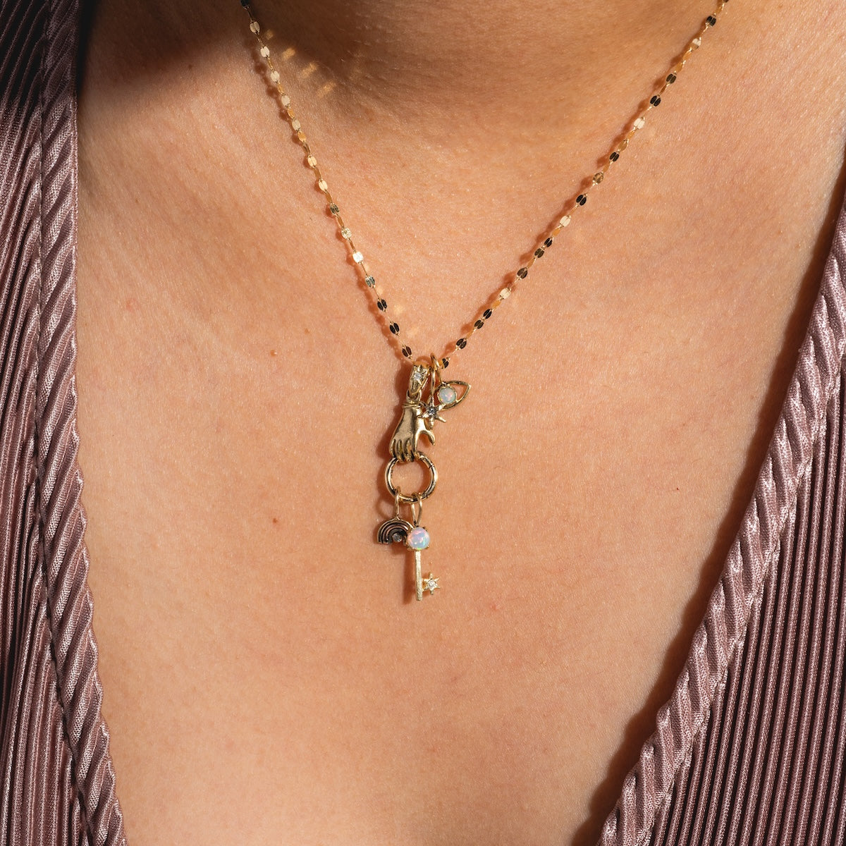 A close-up of the 14k Solid Gold Tiny Charms by Acanthus, featuring gold beads and charms like a key and eye, rests on a person’s neck above a light ribbed mauve top.