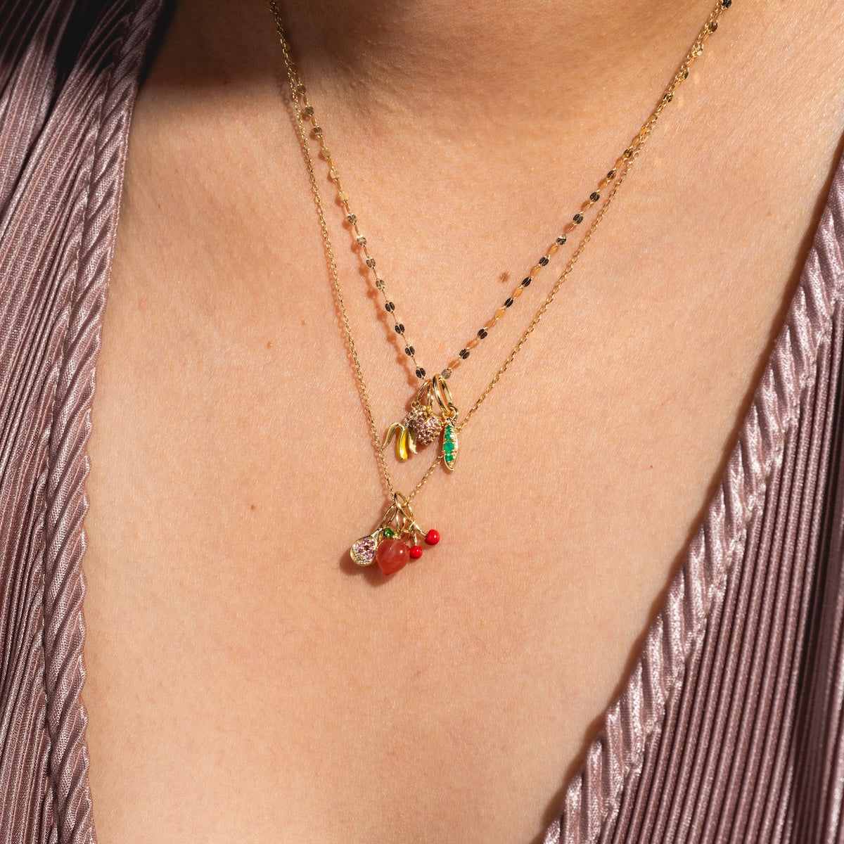 A close-up of a person wearing two delicate 9k yellow gold necklaces, featuring the Solid Gold Foodie Charm and fruit-inspired elements, over a pleated mauve top.