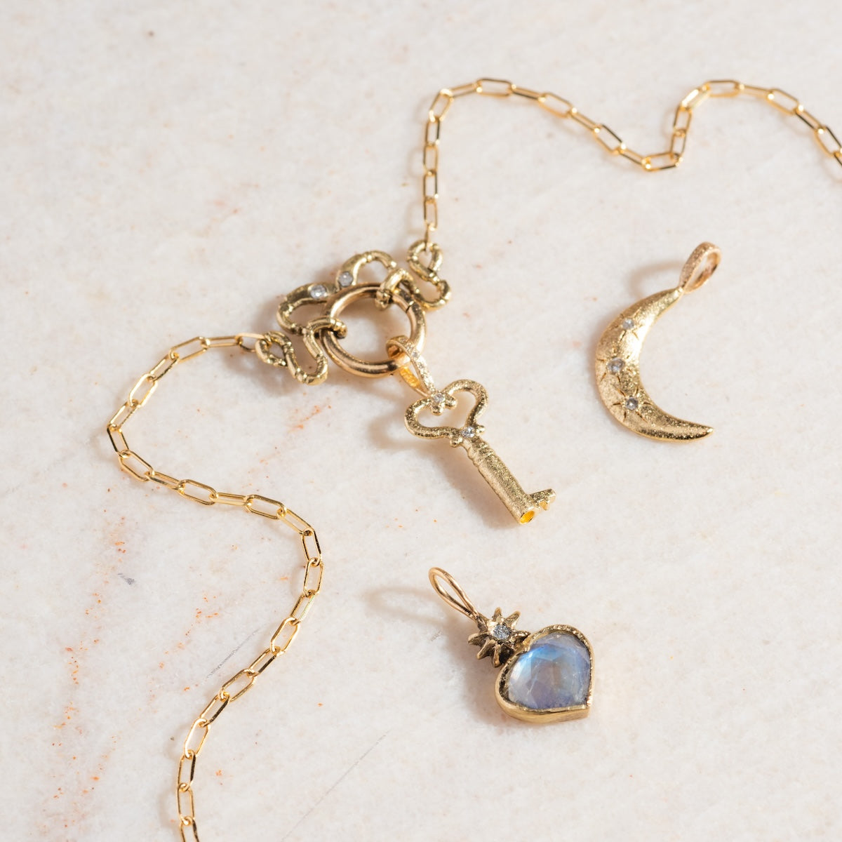 A gold chain necklace with a key-shaped pendant is shown on a light surface, beside a 14k solid yellow gold crescent moon charm and the Guided Heart Pendant Charm featuring a blue gemstone and diamond starburst.