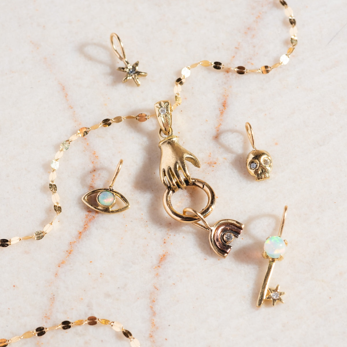 14k Solid Gold Tiny Charms by Acanthus—including a hand, eye with opal, skull, star, and opal bar—are displayed on a light marble surface beside a delicate gold chain.