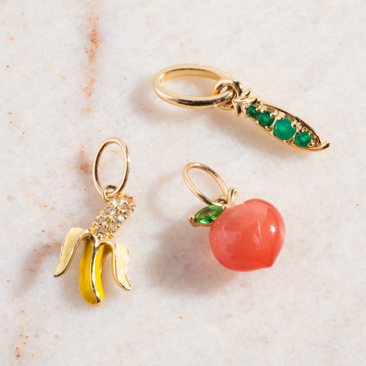Three Solid Gold Foodie Charms rest on marble: a yellow and white banana with crystals, a pea pod with green gems, and a peach with a green leaf—perfect for any fruit charm necklace.
