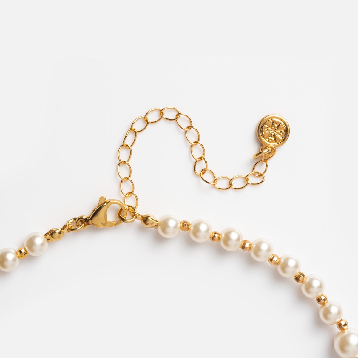 Close-up of the Penelope Necklace, a handmade piece with a gold chain, white pearls, lobster clasp, adjustable extension chain, and a round gold charm with a decorative symbol, displayed on a white background.