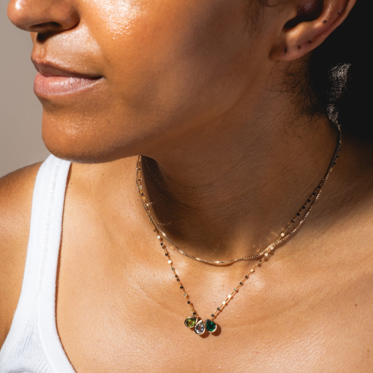 A woman wearing a white tank top and layered gold necklaces, including one featuring the Solid Gold Pear Gemstone Slider Charm, is shown from the shoulders up in soft lighting.