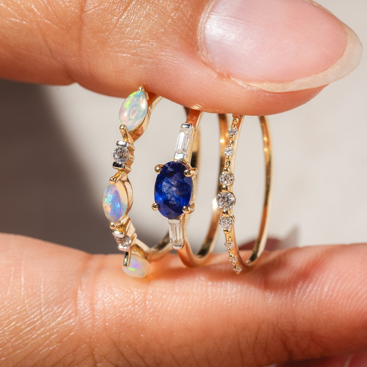 A hand holds three gold rings: a delicate opal and diamond shimmer ring, a blue oval gemstone ring with baguette diamonds, and the Solid Gold Lab Diamond Sparkle Stacking Band with round diamonds.