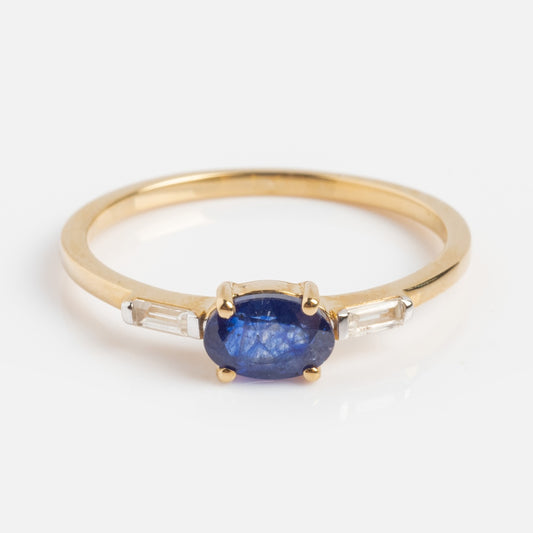 The Solid Gold Classic Sapphire Ring features a 9k solid gold band with an oval fissure-filled blue sapphire center, accented by two baguette-cut white diamonds on each side, set against a white background.